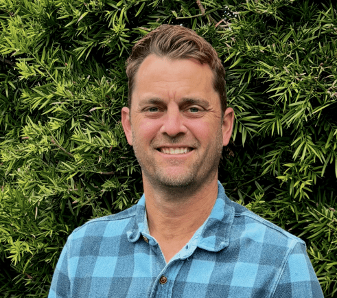 A man in a blue plaid shirt stands smiling in front of lush green foliage.